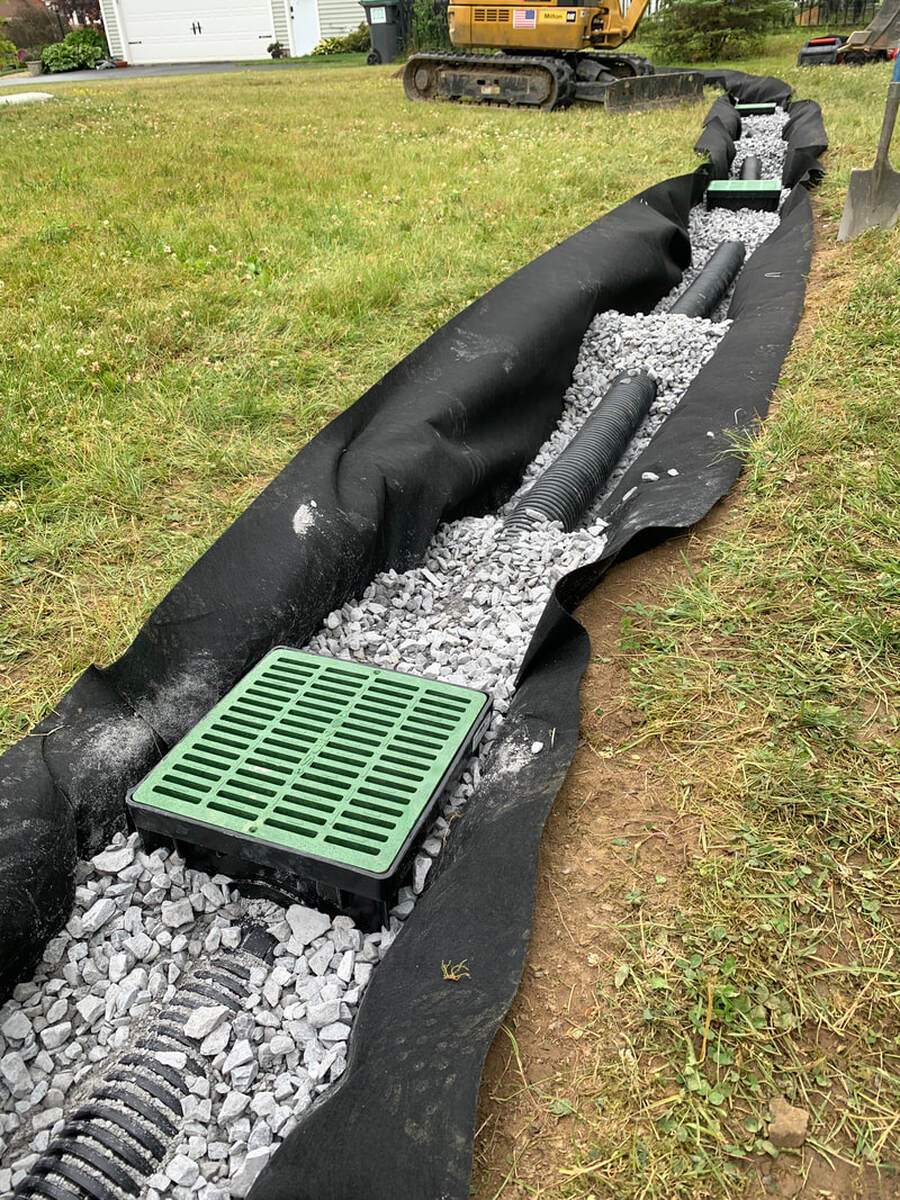 How to make a french drain picture.  Landscape drainage solution for water in the yard.  Building a french drain with catch basins, perforated pipe, and fabric wrapped stone trench.  DIY french drain ideas for a sloped yard or outside of a house.  How to install a french drain diy landscaping idea.